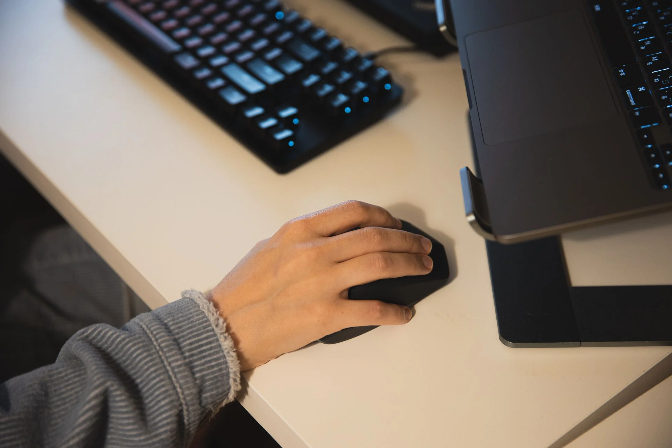 A hand uses a computer mouse beside a laptop and keyboard.
