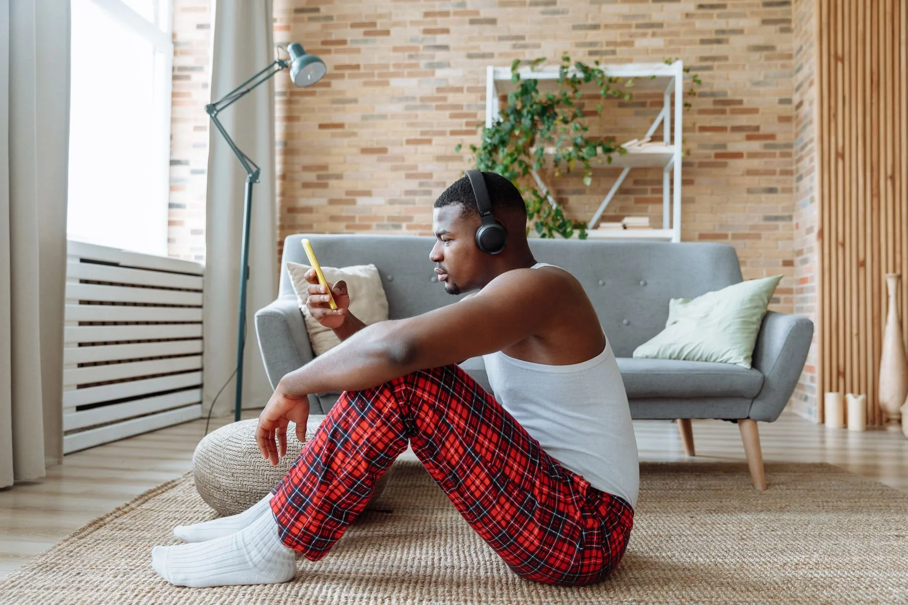 A man wearing headphones looks at his smartphone in a bright living room.