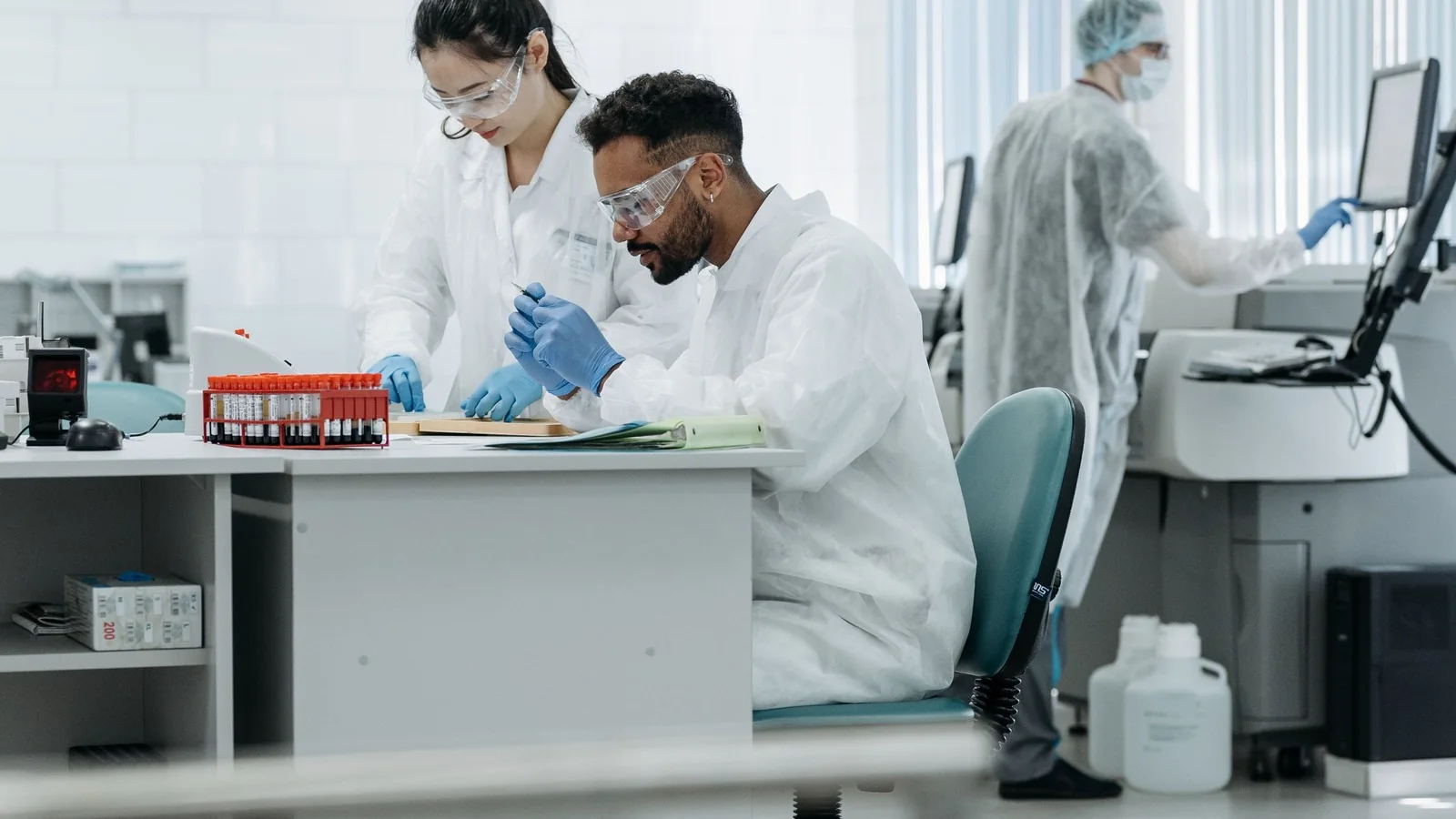 Two researchers examine a sample at a laboratory bench while another works at a computer.